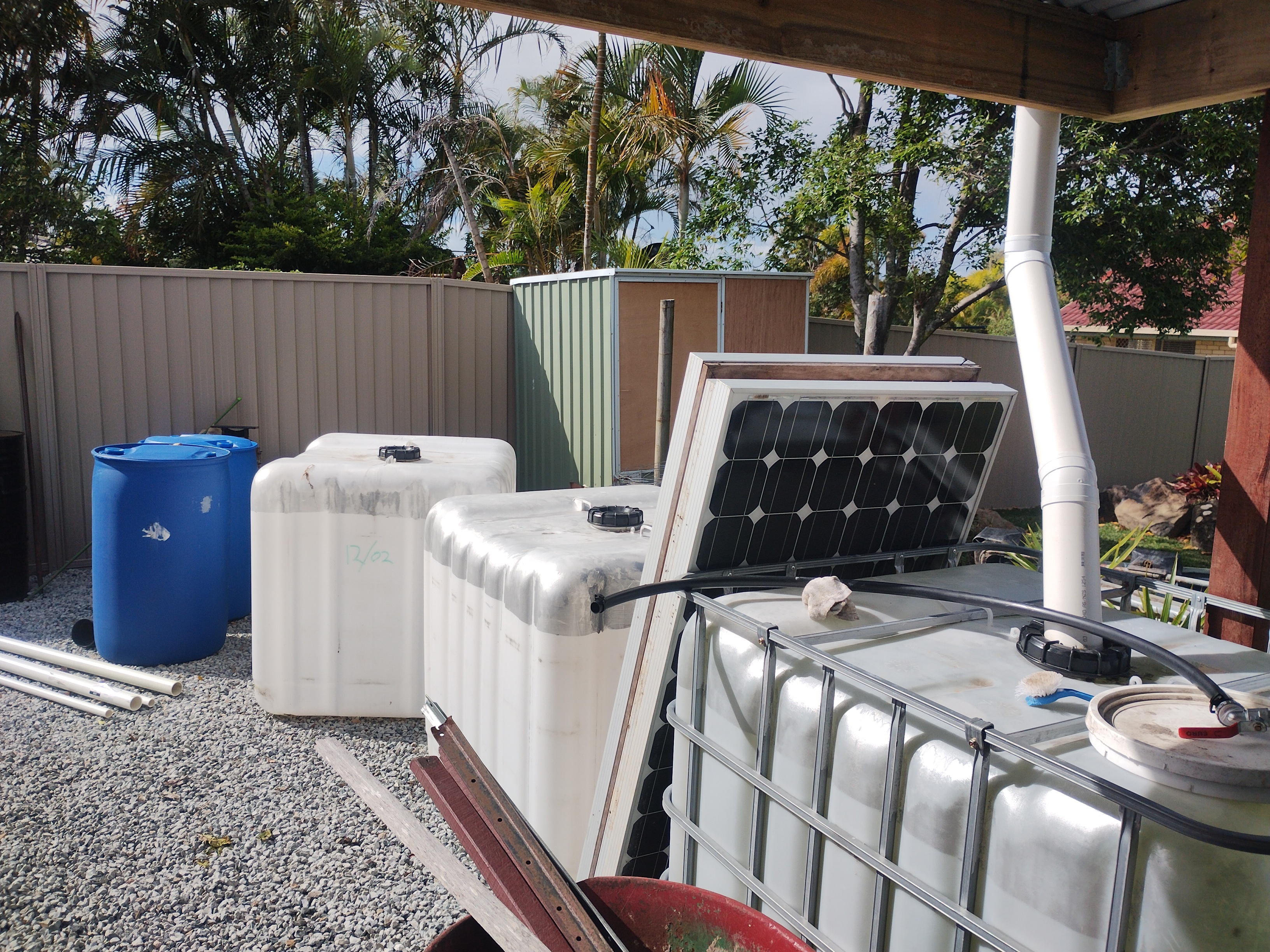 White PVC downpipe from the shanty gutter running into an IBC tank, with a blue drum and other water storage tanks on the gravel beside it, and a solar panel leaning nearby